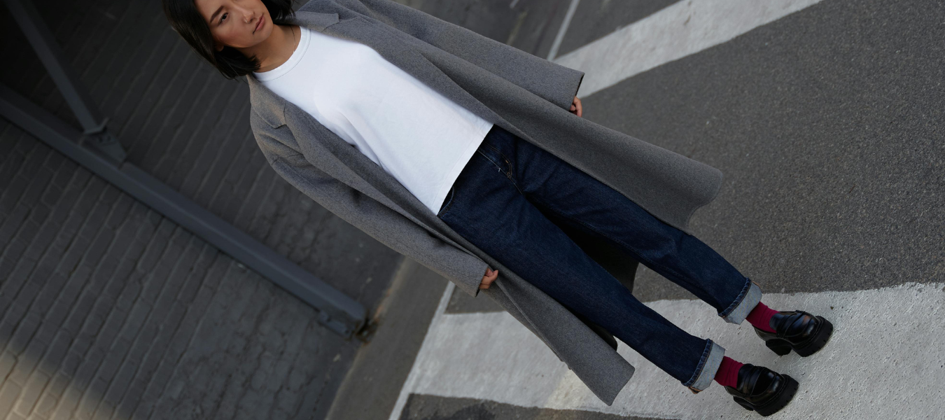 Stylizacja jesienna – szary płaszcz damski, klasyczne jeansy, biała koszulka oraz czarne loafersy z kolorowymi skarpetkami – modne buty na jesień 2025