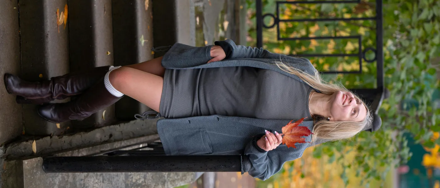 Jesienna stylizacja z szarą dzianinową sukienką i brązowymi kozakami przed kolano, kobieta na schodach z liściem w ręku