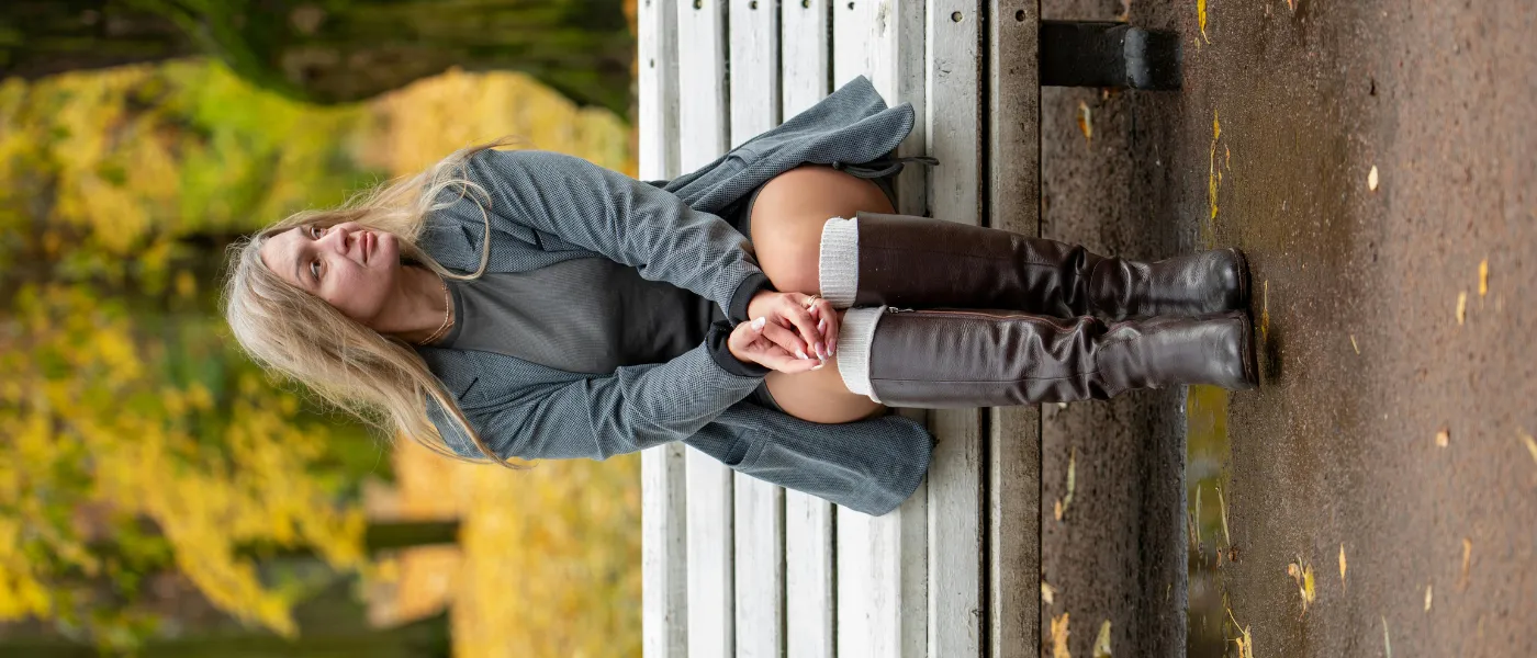 Jesienna stylizacja z długimi brązowymi kozakami, szarą sukienką i kardiganem na ławce w parku