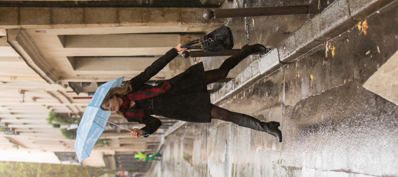 Kobieta z niebieską parasolką w deszczowy dzień, ubrana w czarny płaszcz, czerwony szalik i czarne kozaki – jesienna moda uliczna