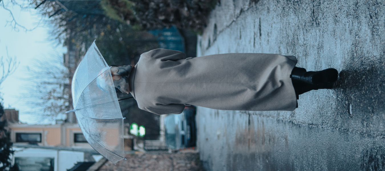 Elegancka kobieta w długim płaszczu idąca mokrym chodnikiem w deszczu, przezroczysty parasol i czarne wodoodporne buty damskie – jesienna stylizacja